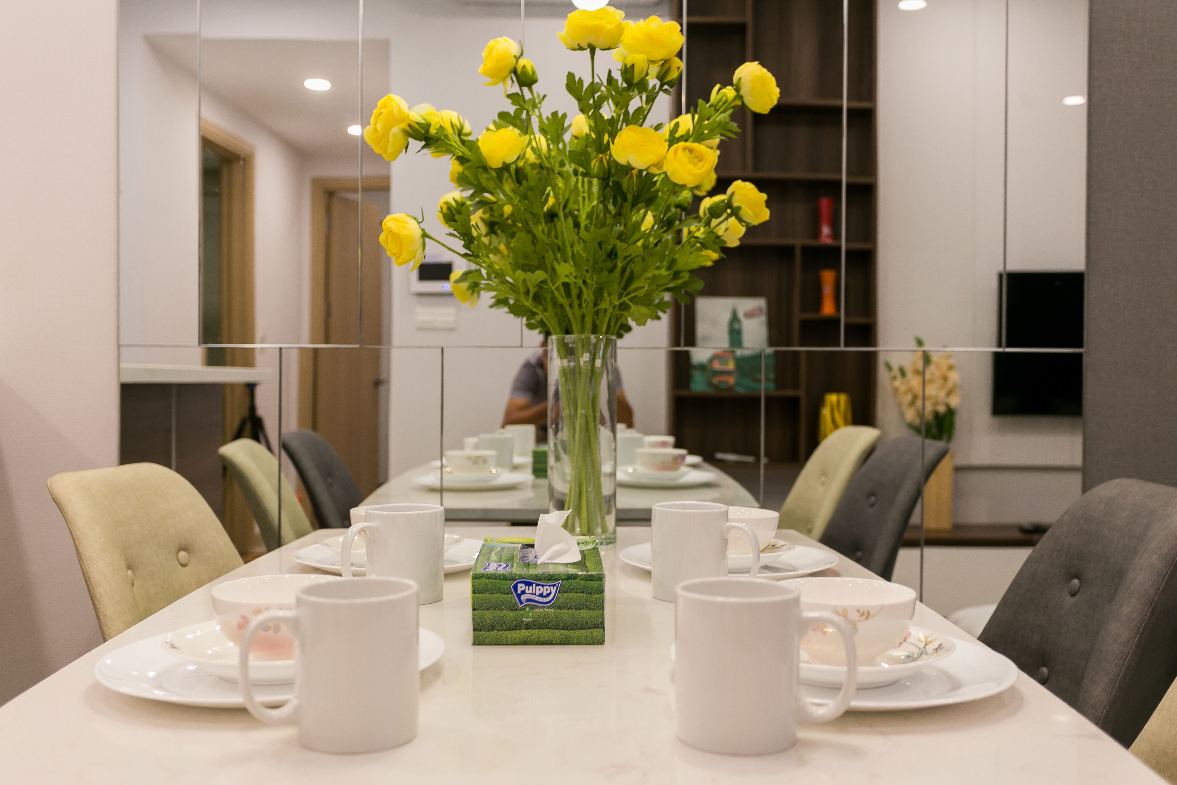 A dining table set with white plates, cups, and a vase of yellow flowers.