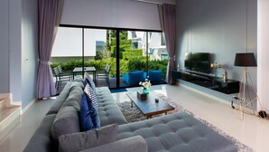 A modern living room with a large sofa, a coffee table, and a TV. There is a balcony with a view of greenery and buildings. - Utopia Naiharn (Rawai)