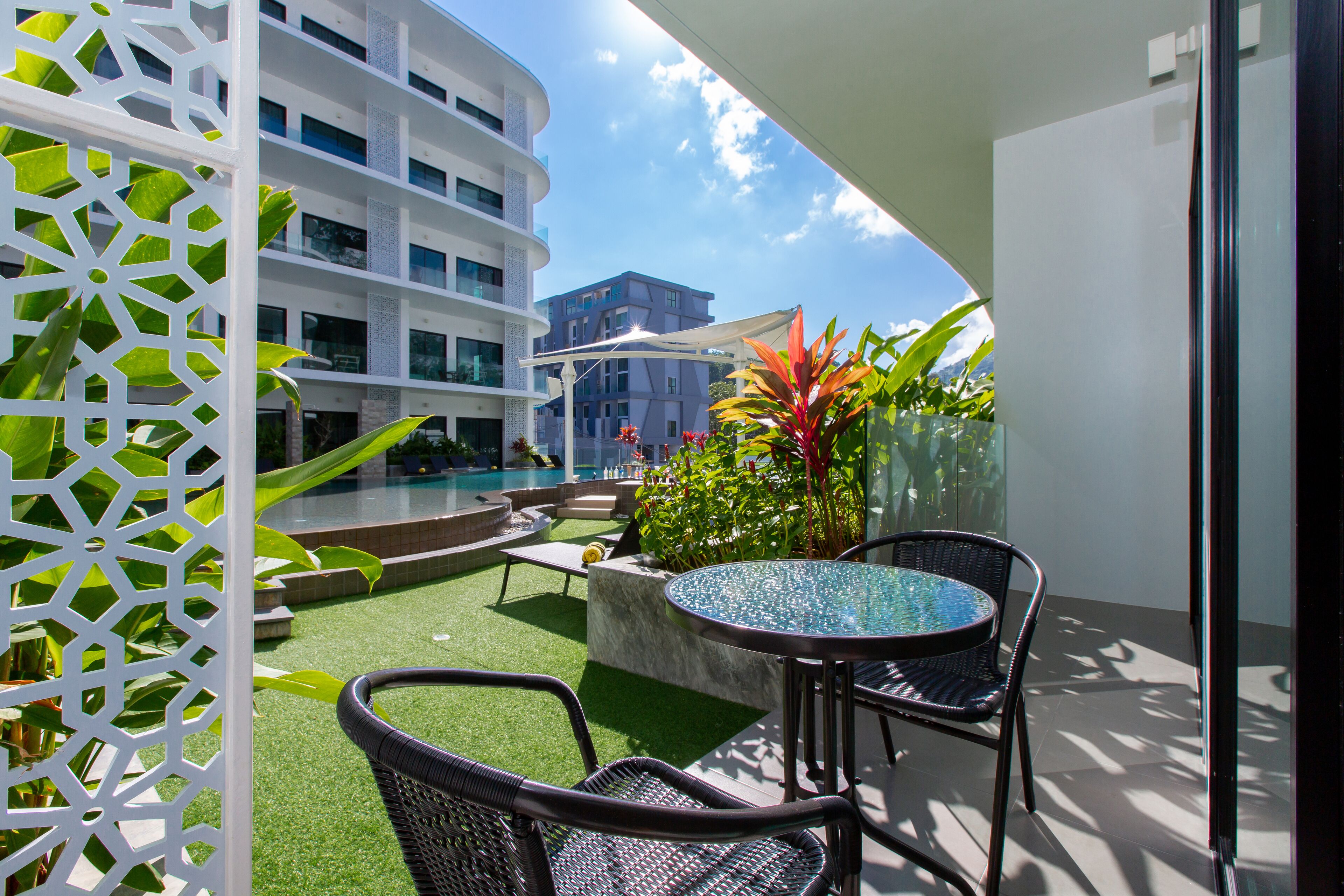 A balcony with artificial grass, a glass table, wicker chairs, and a view of a pool and buildings.