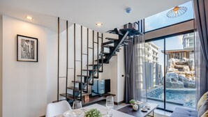 A modern living room with a dining area, a staircase, and a view of a pool and buildings. - Utopia Naiharn (Rawai)