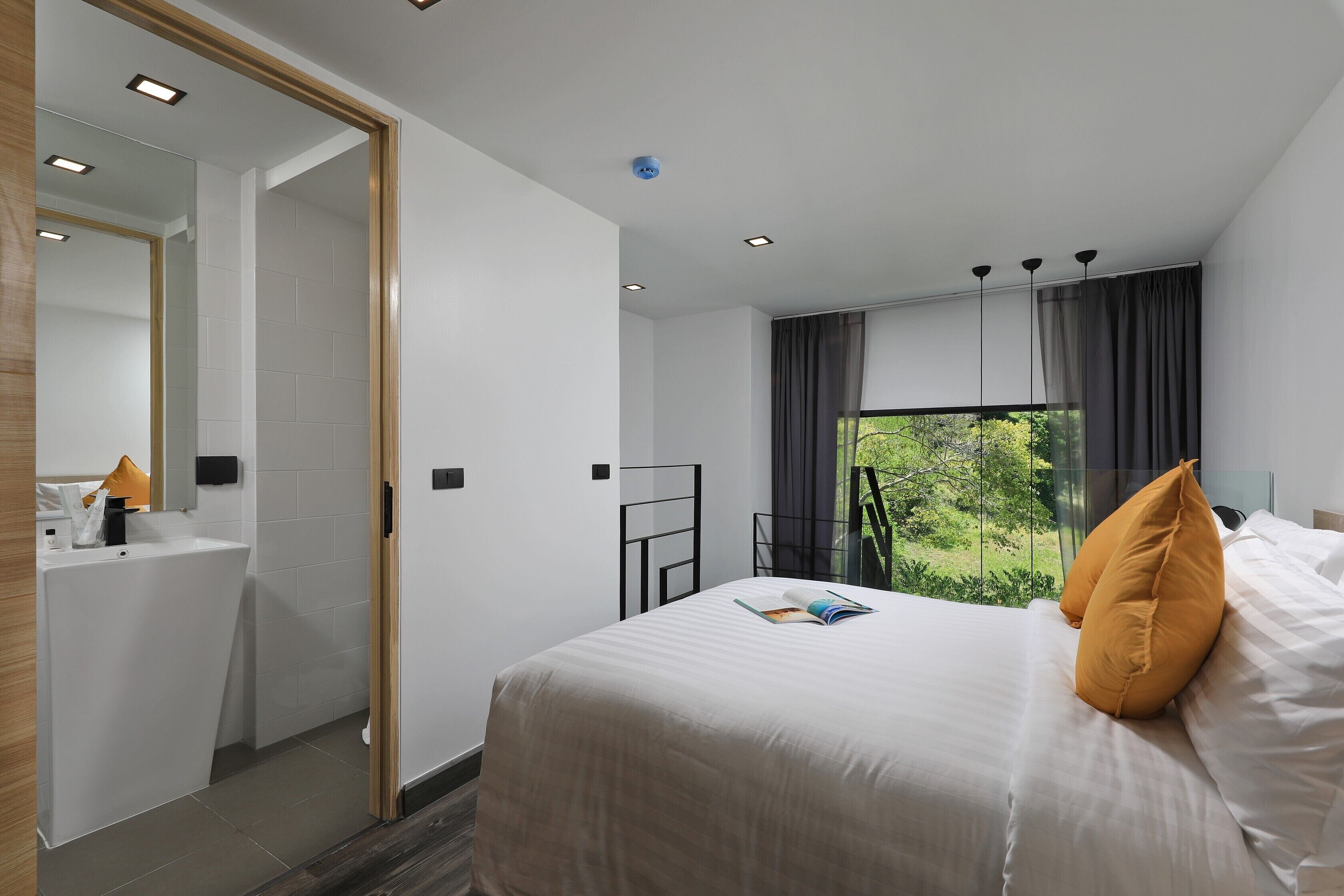 A modern hotel room with a large bed, a bathroom with a sink, and a view of greenery through the window.