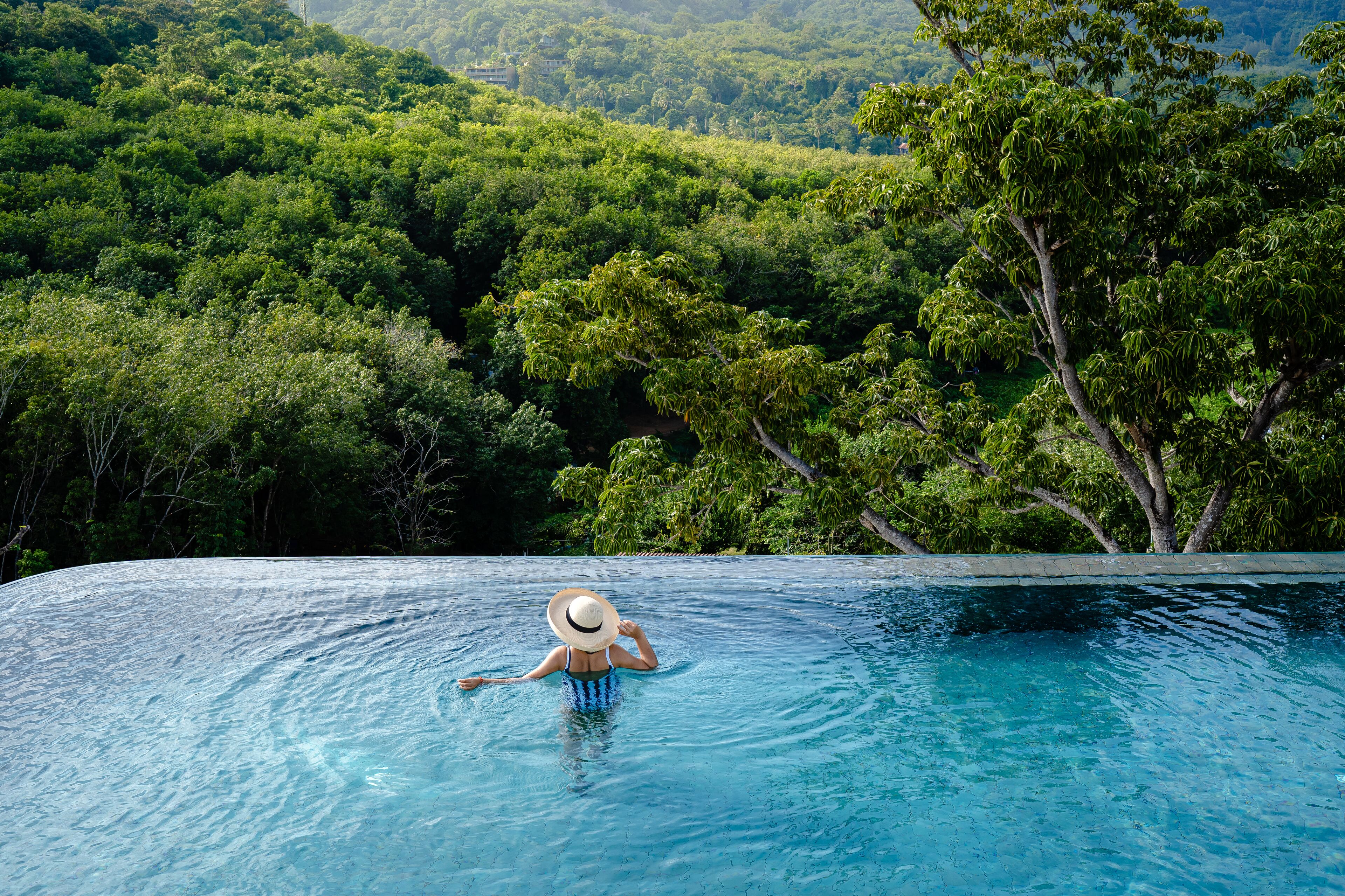 A person relaxing in a clear blue infinity pool with a lush green forest in the background.