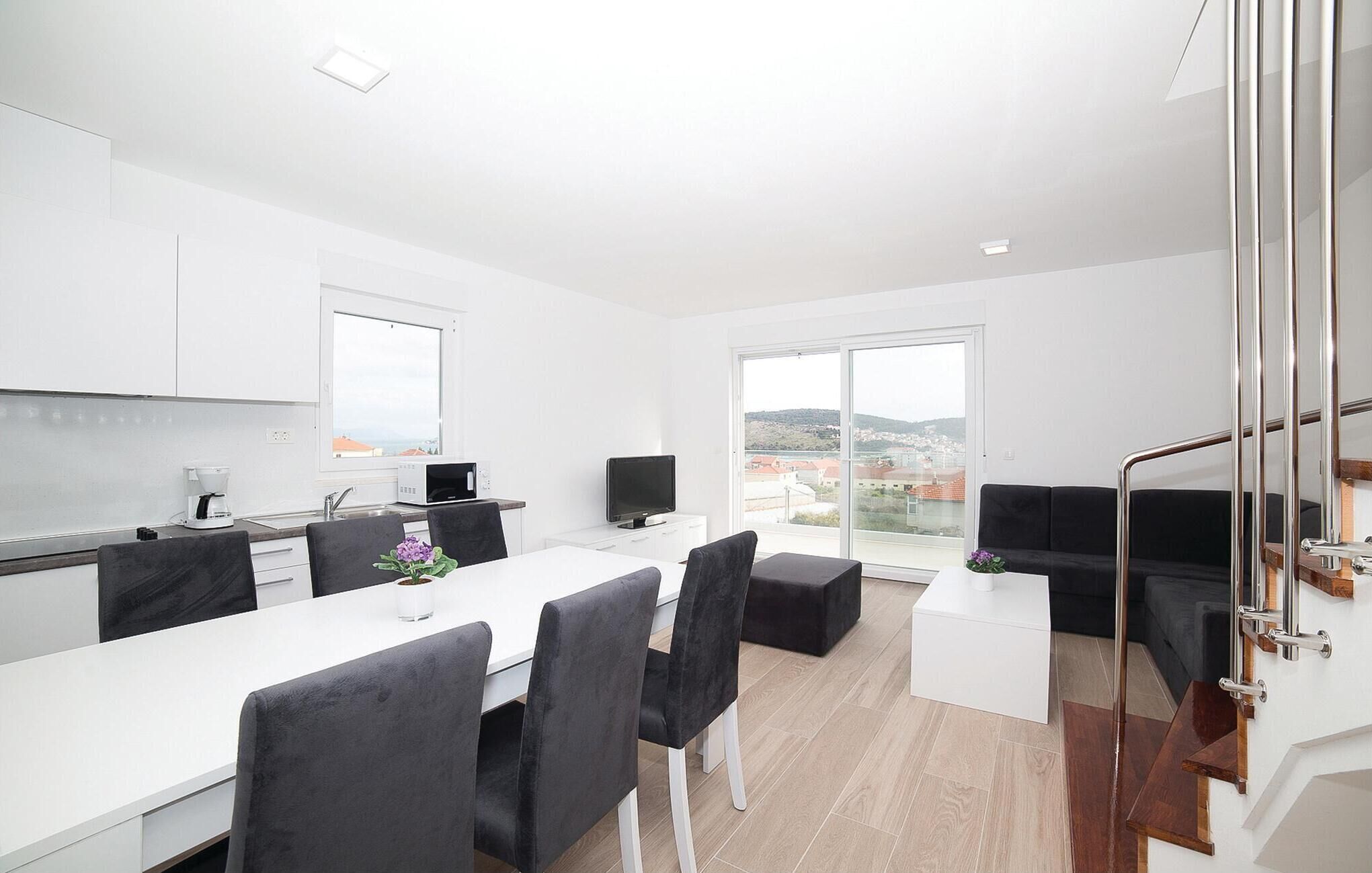A modern kitchen with white cabinets, a dining area with a white table and black chairs, and a large window offering a view of the city.