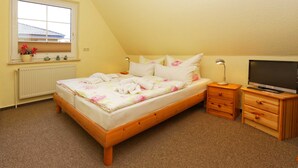 A bedroom with a wooden bed, a window with flowers, a radiator, a TV, and wooden nightstands. - Vacation apartment forest and sea (Loddin)