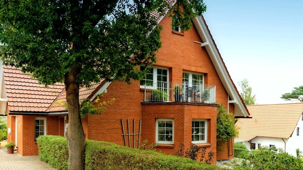 A two-story brick house with a sloped roof, a balcony on the second floor, and a paved driveway. - Vacation apartment forest and sea (Loddin)