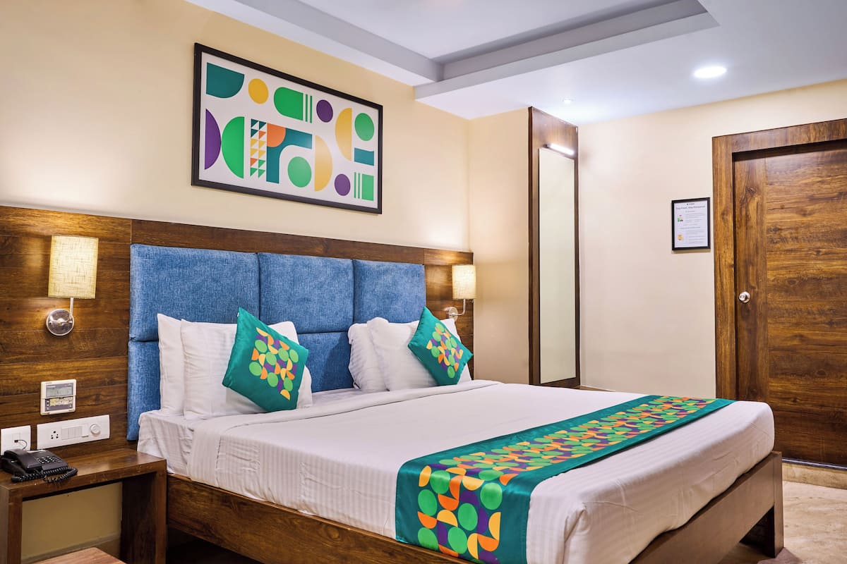 Een hotelkamer met een houten hoofdeinde, een bed met wit beddengoed, een kleurrijk plaid, een bedlampje en een aan de muur bevestigd kunstwerk.