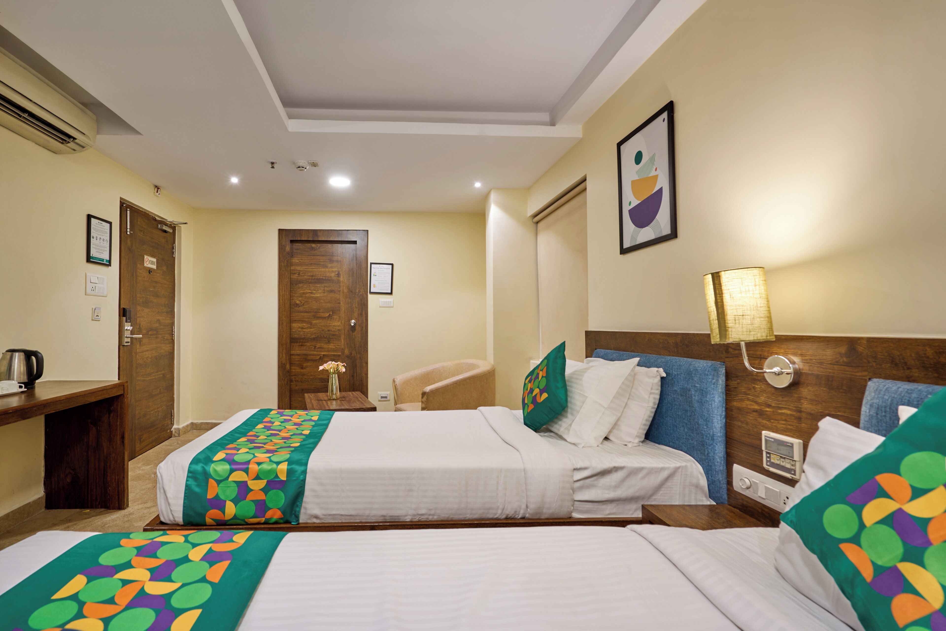 A hotel room with two beds, a desk, a chair, a bedside table, a lamp, and a wall-mounted clock.