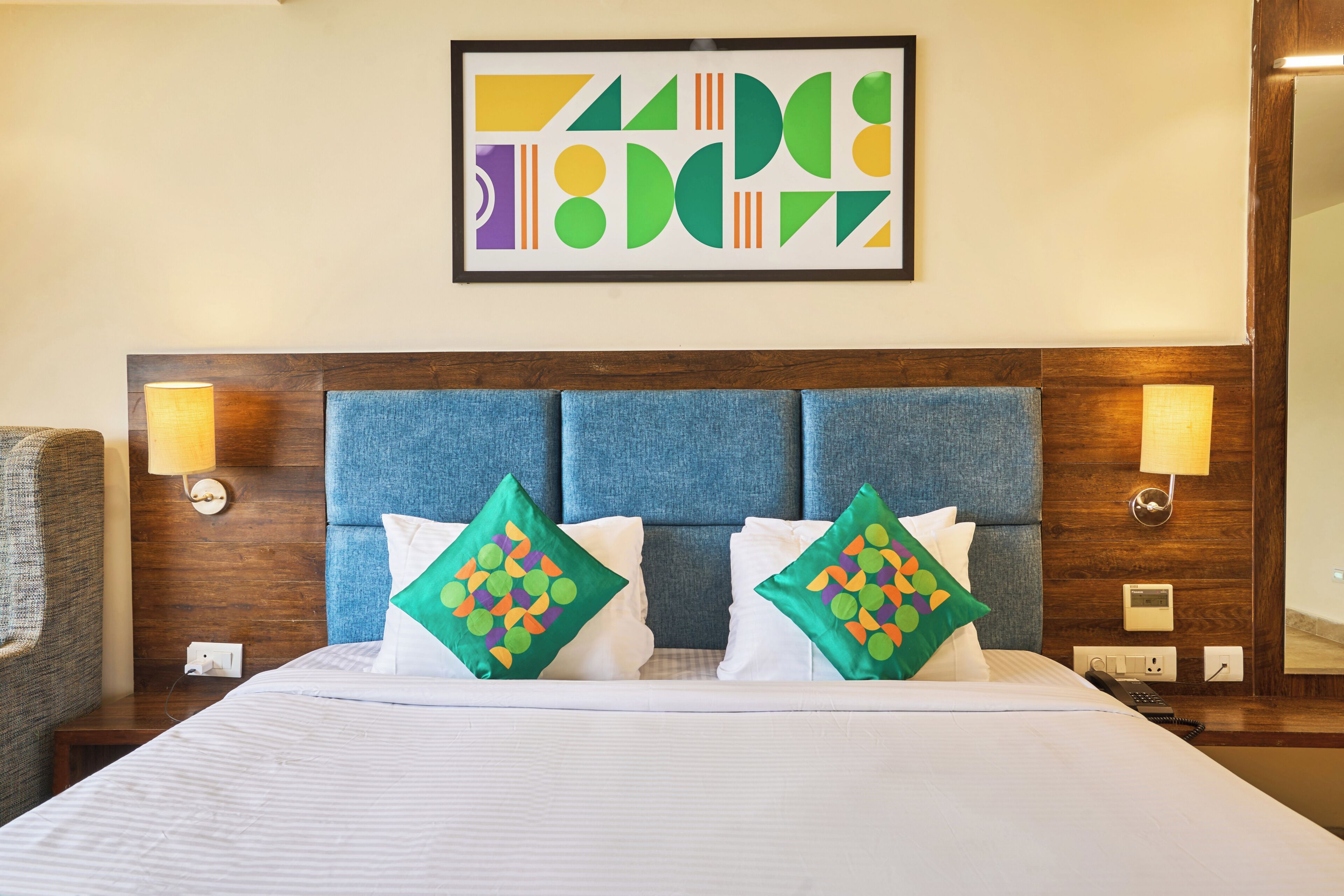 A hotel room with a large bed, two decorative pillows, a headboard, wall-mounted lamps, and a framed abstract artwork.