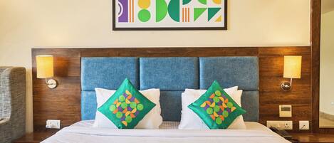 A hotel room with a large bed, two decorative pillows, a headboard, wall-mounted lamps, and a framed abstract artwork.
