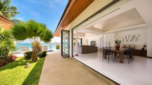 Una casa moderna con un ventanal de vidrio que da a un patio con piscina, zona de comedor y sala de estar con vista al mar.