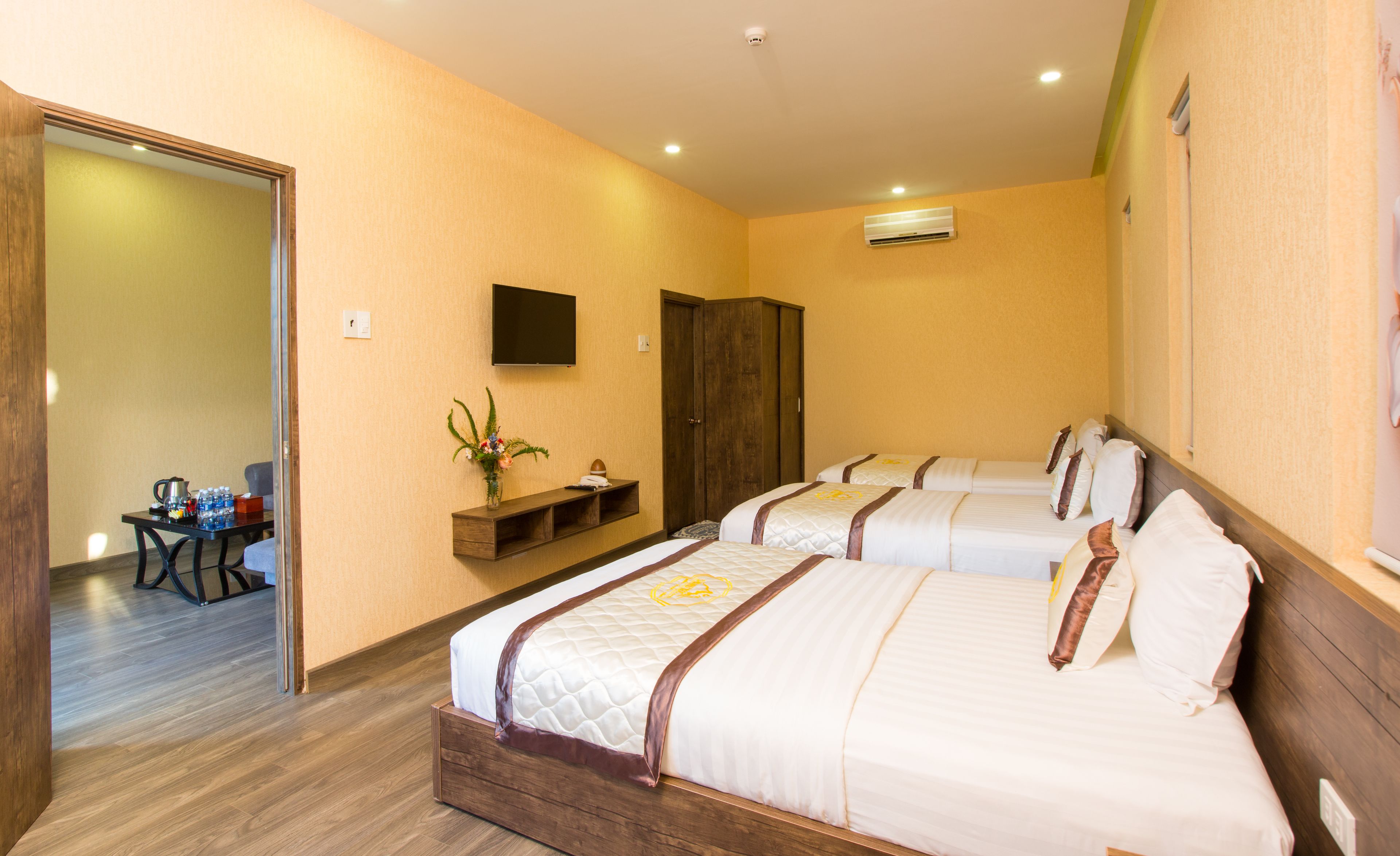 Quarto de hotel com duas camas, uma televisão, guarda-roupa de madeira, uma pequena mesa com um vaso de flores e um banheiro com uma toalha pendurada na parede.