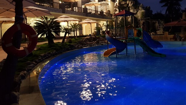 Un espace piscine nocturne doté de toboggans et de parasols, entouré d’un bâtiment à plusieurs niveaux et offrant des espaces de détente en plein air.