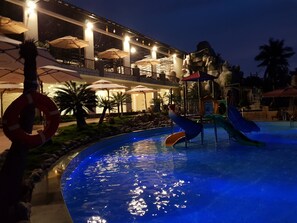 Un espace piscine nocturne doté de toboggans et de parasols, entouré d’un bâtiment à plusieurs niveaux et offrant des espaces de détente en plein air.