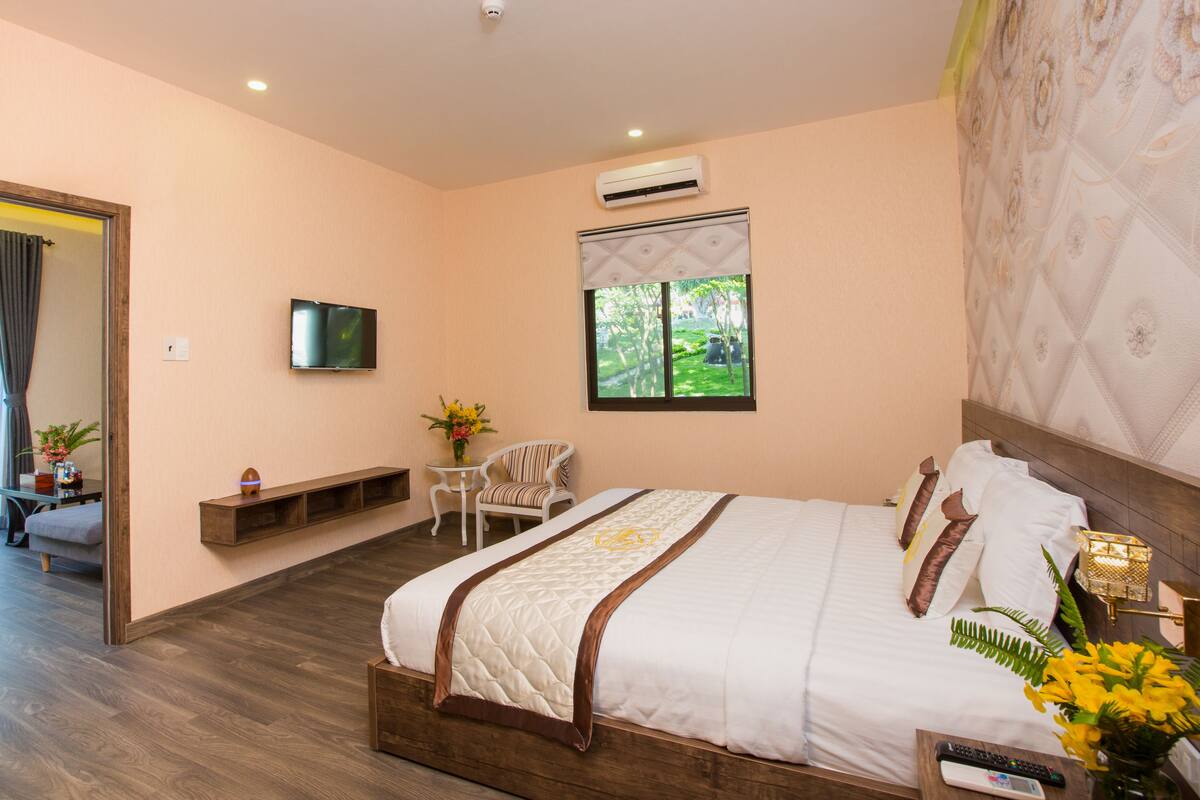 Habitación de hotel con una cama grande, un televisor montado en la pared, una ventana con vistas a zonas verdes, una mesita con una silla y un equipo de aire acondicionado.