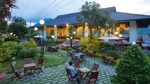 Un espace de restauration en plein air, avec des tables et des chaises, entouré de verdure et bordé d’un bâtiment dont le toit est recouvert de tuiles.