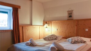 Een kamer met twee bedden, houten lambrisering, een raam met gordijnen, een wandlamp en een bord met een bijen symbool.
