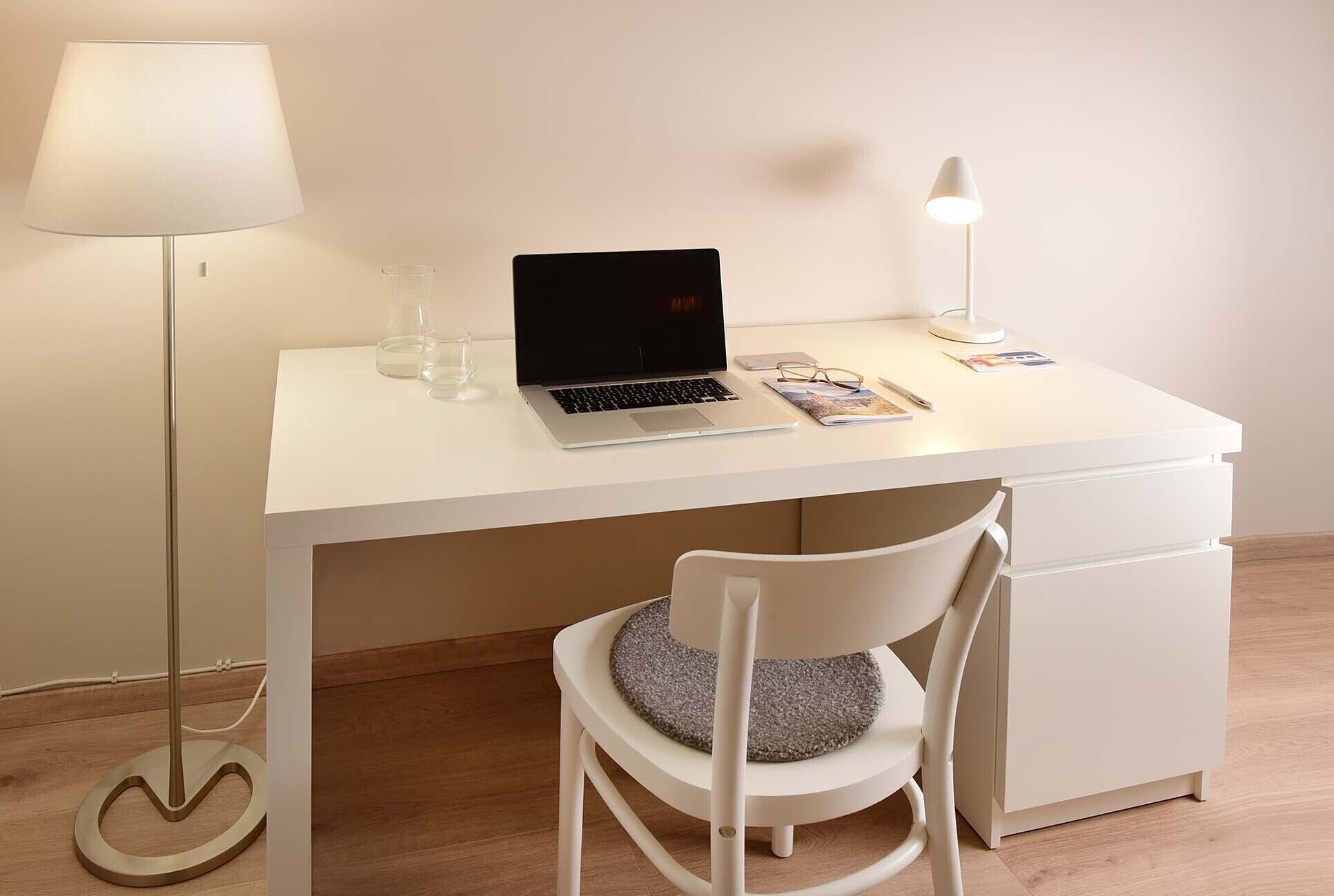 A white desk with a laptop, a lamp, and a chair with a cushion.