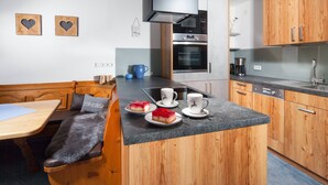 A compact kitchen with wooden cabinets, a built-in oven, and a dining area with a bench and table. - Ferienwohnung 2 bis 2 Personen, 52 qm, Obergeschoss, 1 separates Schlafzimmer, Balkon (Schönau am Königssee)