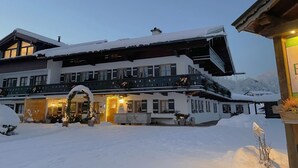 A large, multi-story building with a snow-covered exterior, balconies with railings, and a lit entrance. - Ferienwohnung 2 bis 2 Personen, 52 qm, Obergeschoss, 1 separates Schlafzimmer, Balkon (Schönau am Königssee)