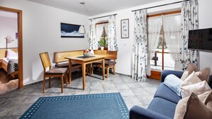 A living room with a sofa, a dining table with chairs, a TV, and a window with curtains. - Ferienwohnung 2 bis 2 Personen, 52 qm, Obergeschoss, 1 separates Schlafzimmer, Balkon (Schönau am Königssee)