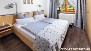 A neatly made bed with checkered and patterned bedding, a bedside table with a lamp, and a window with curtains. - Ferienwohnung 2 bis 2 Personen, 52 qm, Obergeschoss, 1 separates Schlafzimmer, Balkon (Schönau am Königssee)