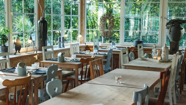 Un comedor con mesas y sillas de madera, ventanas amplias con vistas a zonas verdes y diferentes tipos de mesas dispuestas.