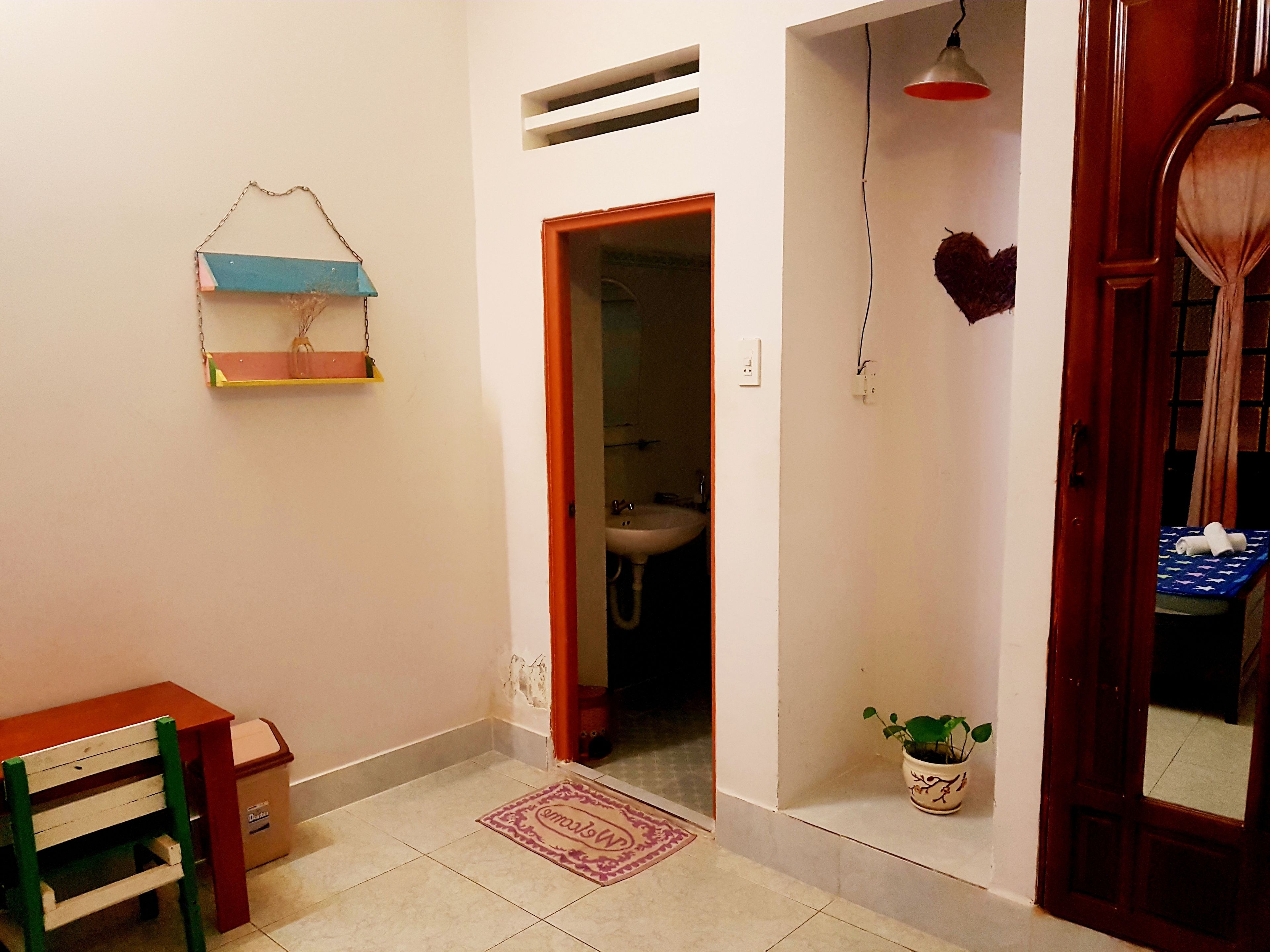Un pasillo estrecho con una puerta de madera, una pequeña mesa con una silla, una lámpara colgante y una planta en maceta.