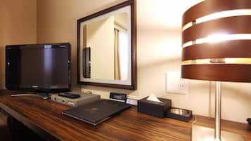 Un bureau dans une chambre d’hôtel, avec un téléviseur à écran plat, une télécommande, un livre, une lampe, une boîte de mouchoirs et un petit bol.