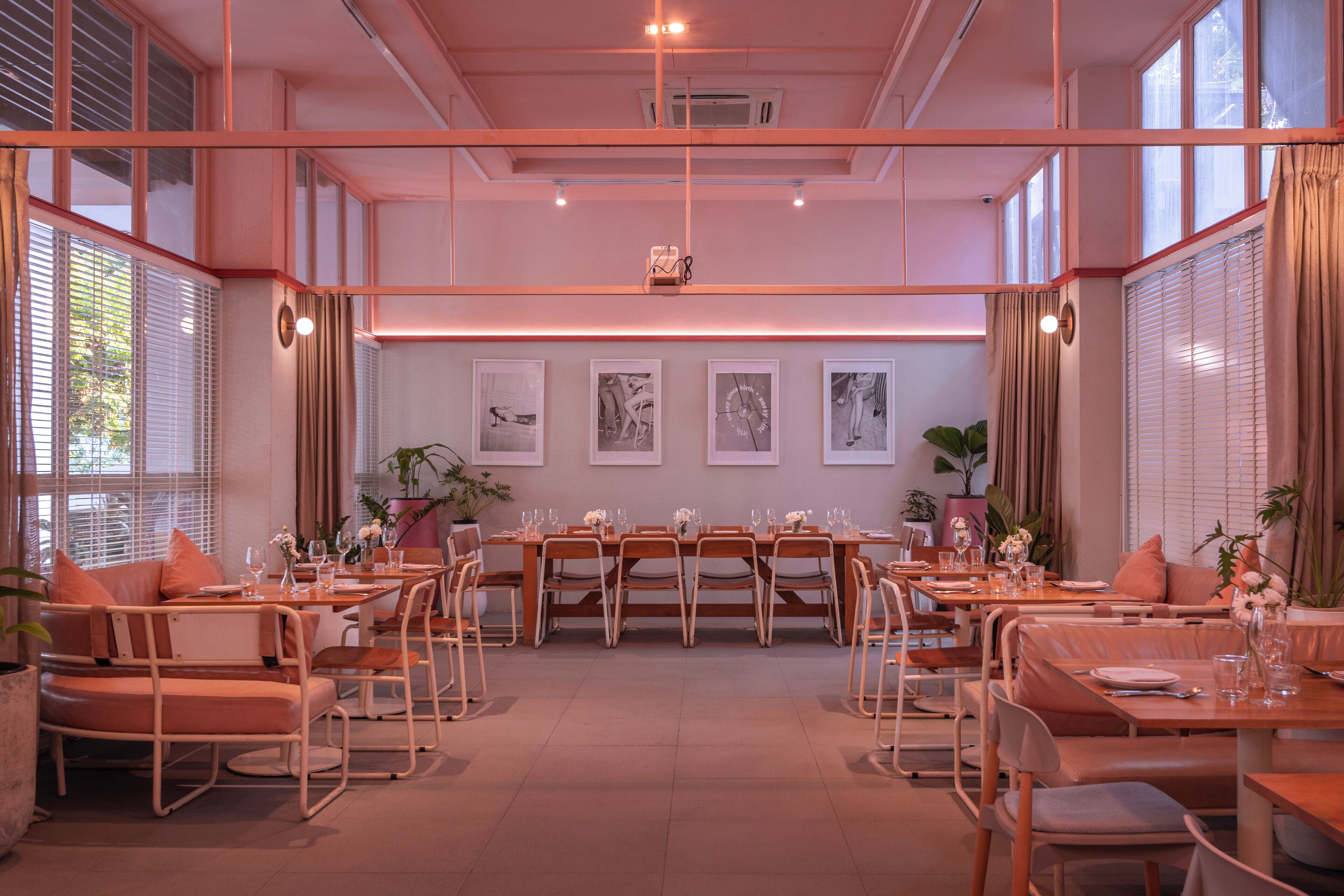 A dining area with multiple tables and chairs, large windows with blinds, and framed artwork on the walls.