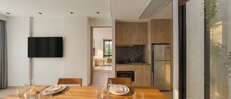 Un comedor moderno con una mesa de madera dispuesta para cuatro comensales, un televisor de pantalla plana montado en la pared y una cocina visible a través de una puerta abierta.