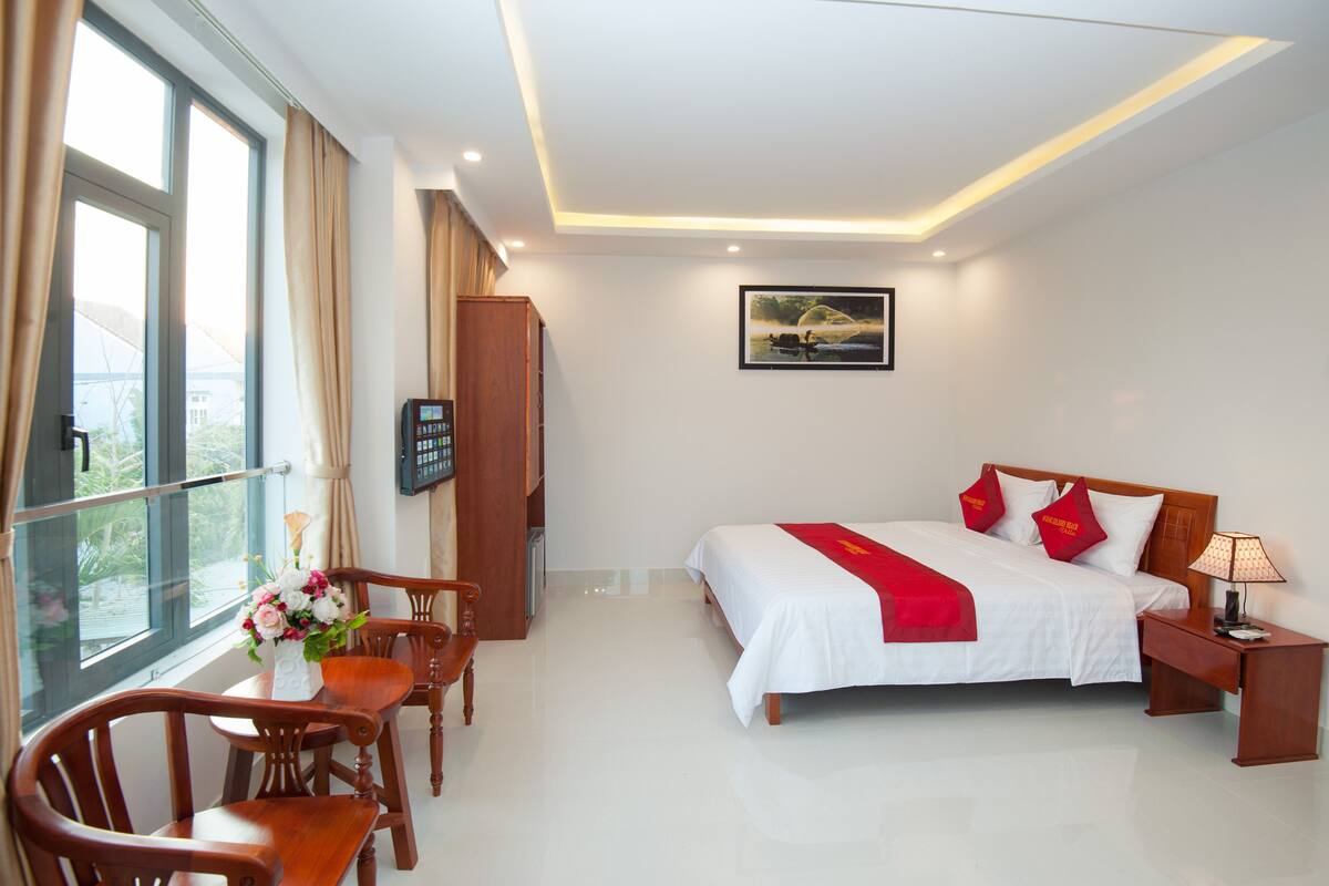 A hotel room with a bed, a desk, a chair, a window with curtains, and a painting on the wall.