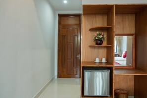Habitación de hotel con puerta de madera, un refrigerador pequeño, repisa con un jarrón de flores y un espejo que refleja parte de la habitación.