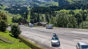Un parking rempli de véhicules, un bâtiment doté d’un toit en pente et, au loin, un paysage montagneux.