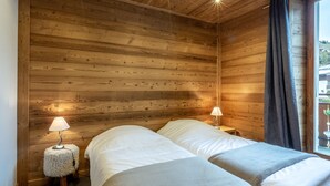 Ein Zimmer in einer Holzhütte mit zwei Betten, einem kleinen Hocker und einem Fenster mit Vorhängen.