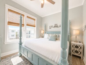 A bedroom with a four-poster bed, a nightstand with a lamp, and a view of buildings through the window.