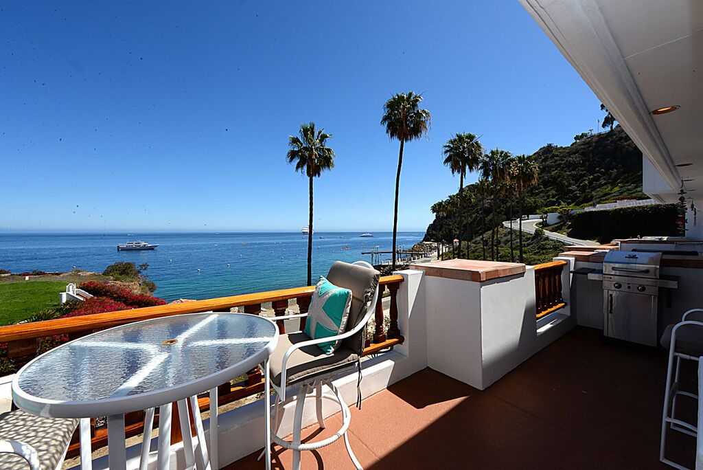 A balcony with a table and chairs, a grill, and a view of the ocean and palm trees.