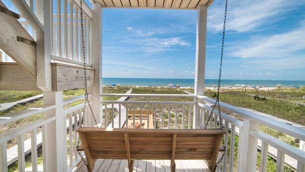 Une chaise suspendue en bois, installée sur une terrasse en bois dotée de rampes blanches, d’où l’on peut admirer une plage et l’océan.