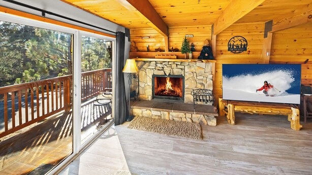 Interior acogedor de una cabaña con chimenea de piedra, televisor montado en la pared y balcón con mobiliario de exterior.