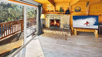 A cozy cabin interior with a stone fireplace, a TV mounted on the wall, and a balcony with outdoor seating.