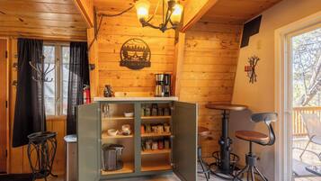A cozy cabin interior with a wooden ceiling, a chandelier, a coffee station, and a dining area with a table and chairs.