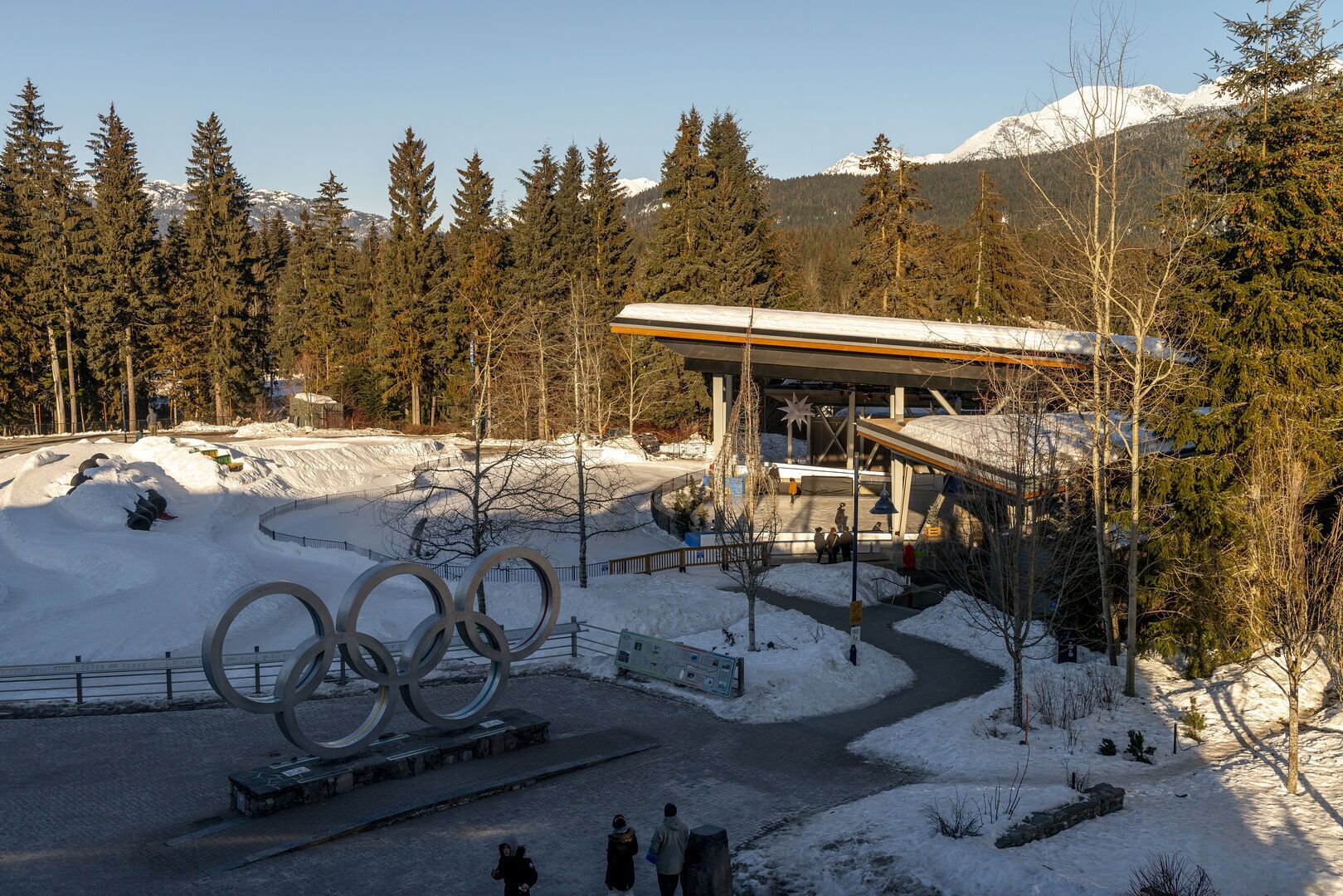 Sneklædt område med en stor skulptur af de olympiske ringe, et overdækket siddeområde og et bjerg i baggrunden.