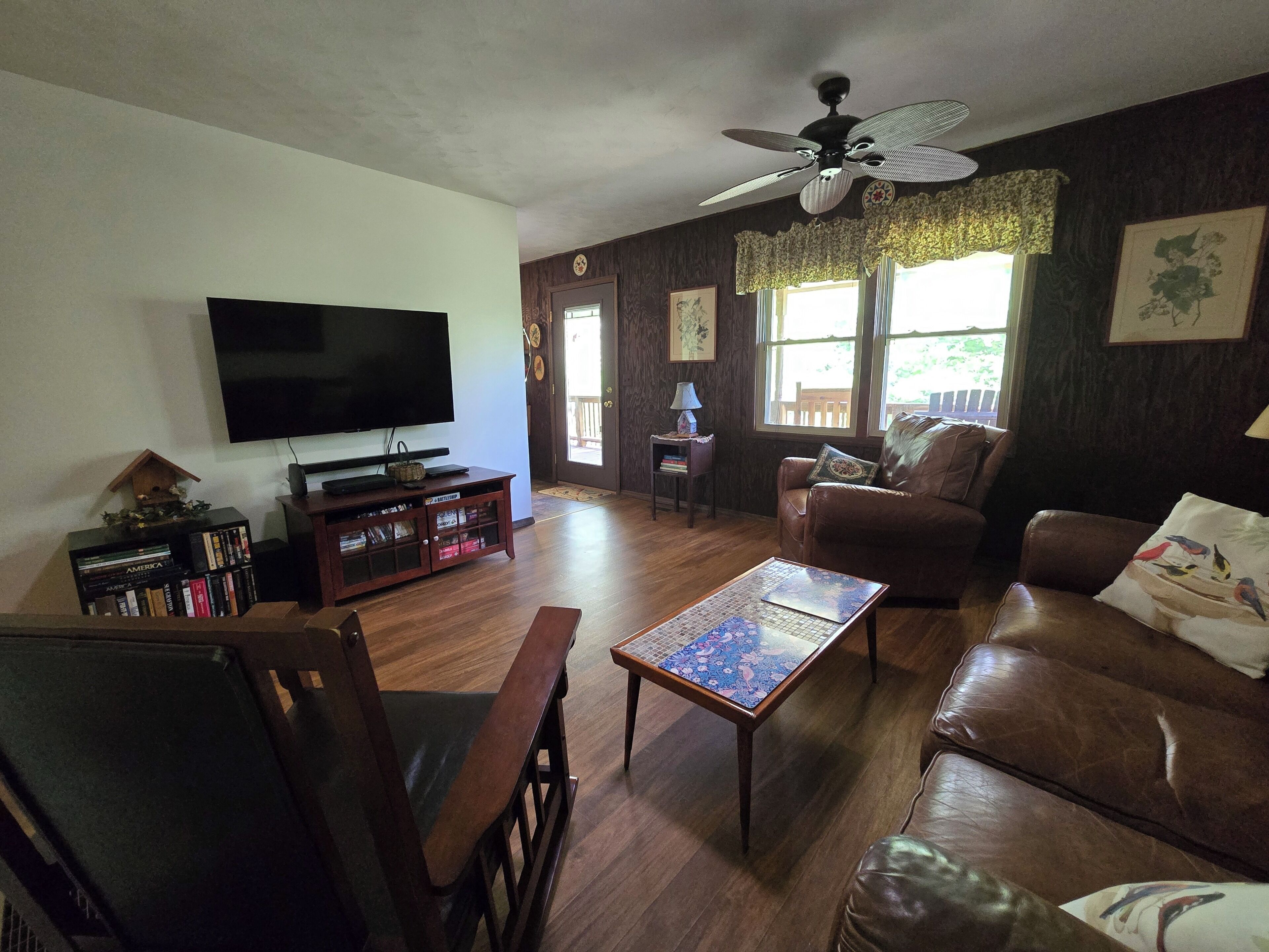 A living room with a television, wooden furniture, a ceiling fan, and large windows.