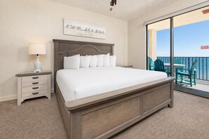 A bedroom with a large bed, a nightstand with drawers, a lamp, and a view of a balcony with chairs.