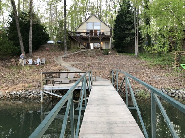Eine Holzterrasse führt zu einem Haus mit Balkon, umgeben von Bäumen und einem kleinen Teich.