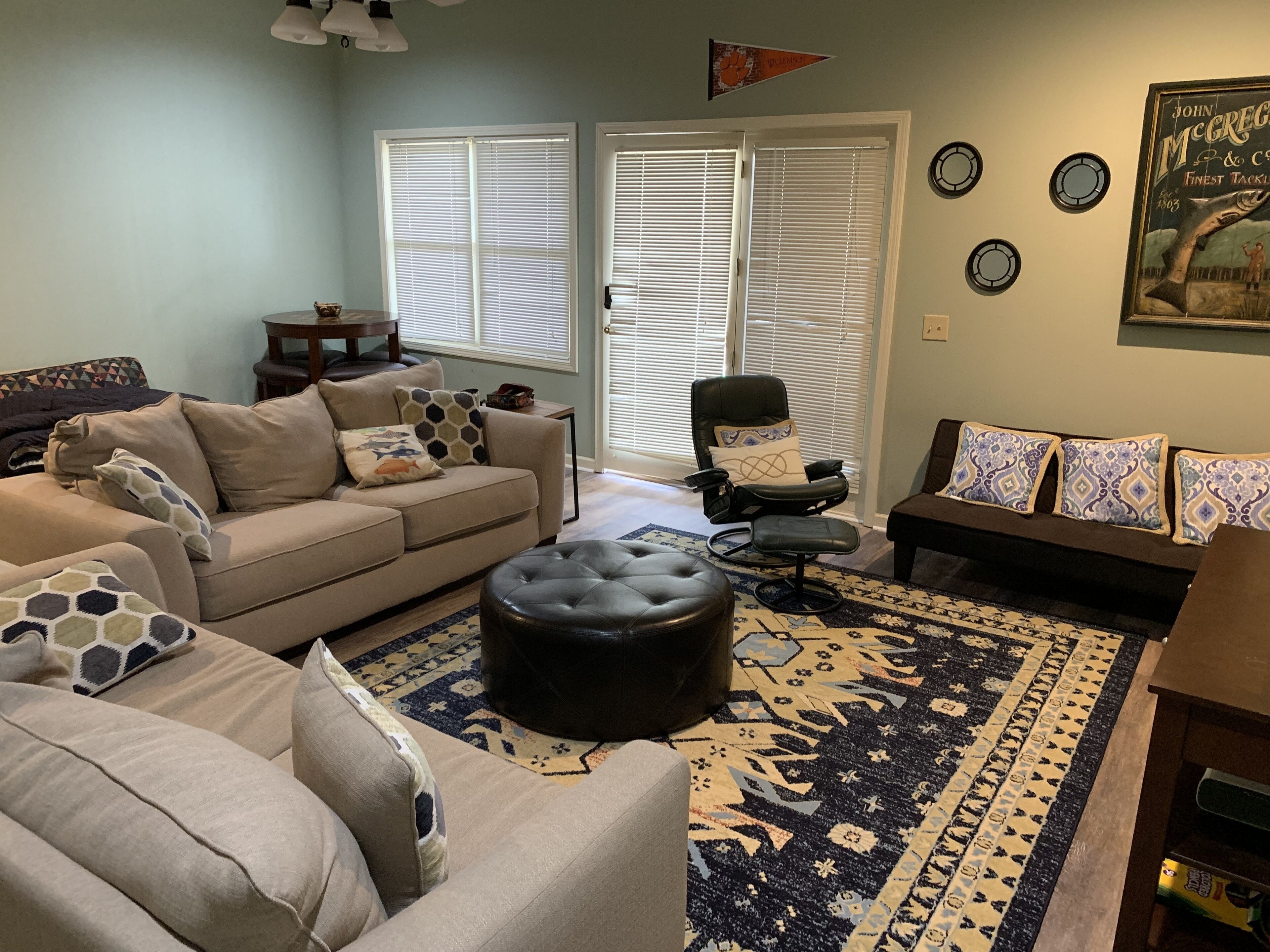 A living room with a beige sofa, a black ottoman, a black chair, and a patterned rug. There are two windows with blinds, a door, and a wall with circular decorations.