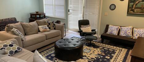 A living room with a beige sofa, a black ottoman, a black chair, and a patterned rug. There are two windows with blinds, a door, and a wall with circular decorations.