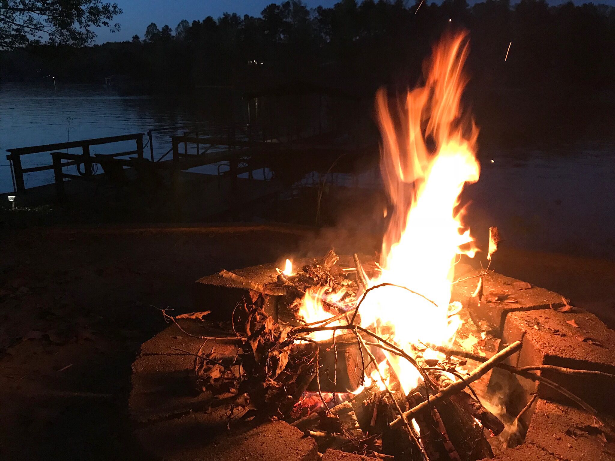 Un fuoco da campo con tronchi e rami, circondato da una recinzione in legno e un molo che si estende sull'acqua.