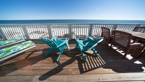 Une terrasse en bois avec deux fauteuils Adirondack turquoise et une table en bois, donnant sur une plage bordée d’une clôture blanche.
