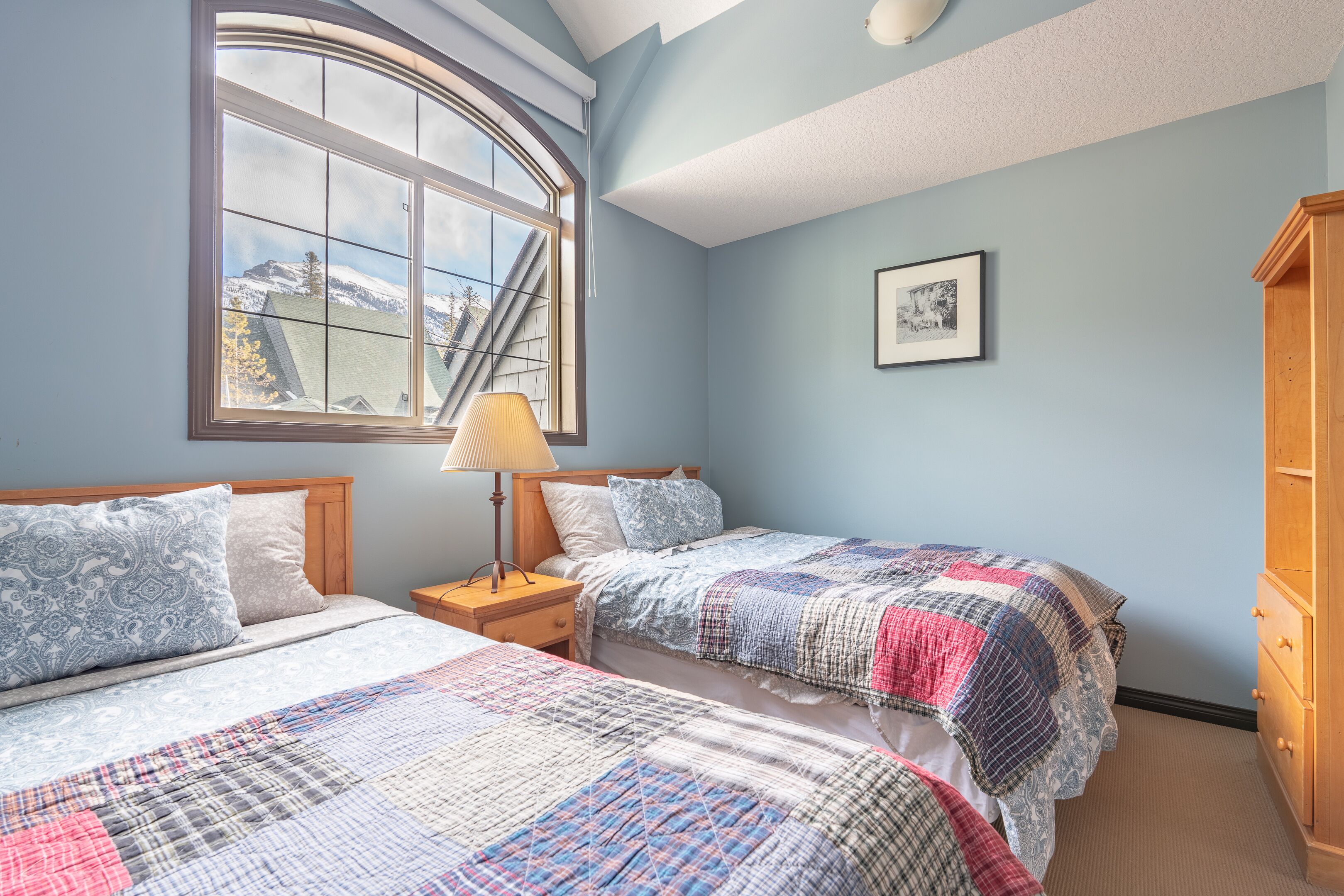 Une chambre avec deux lits, une grande fenêtre donnant sur un beau paysage, une table de chevet avec une lampe et une commode en bois.