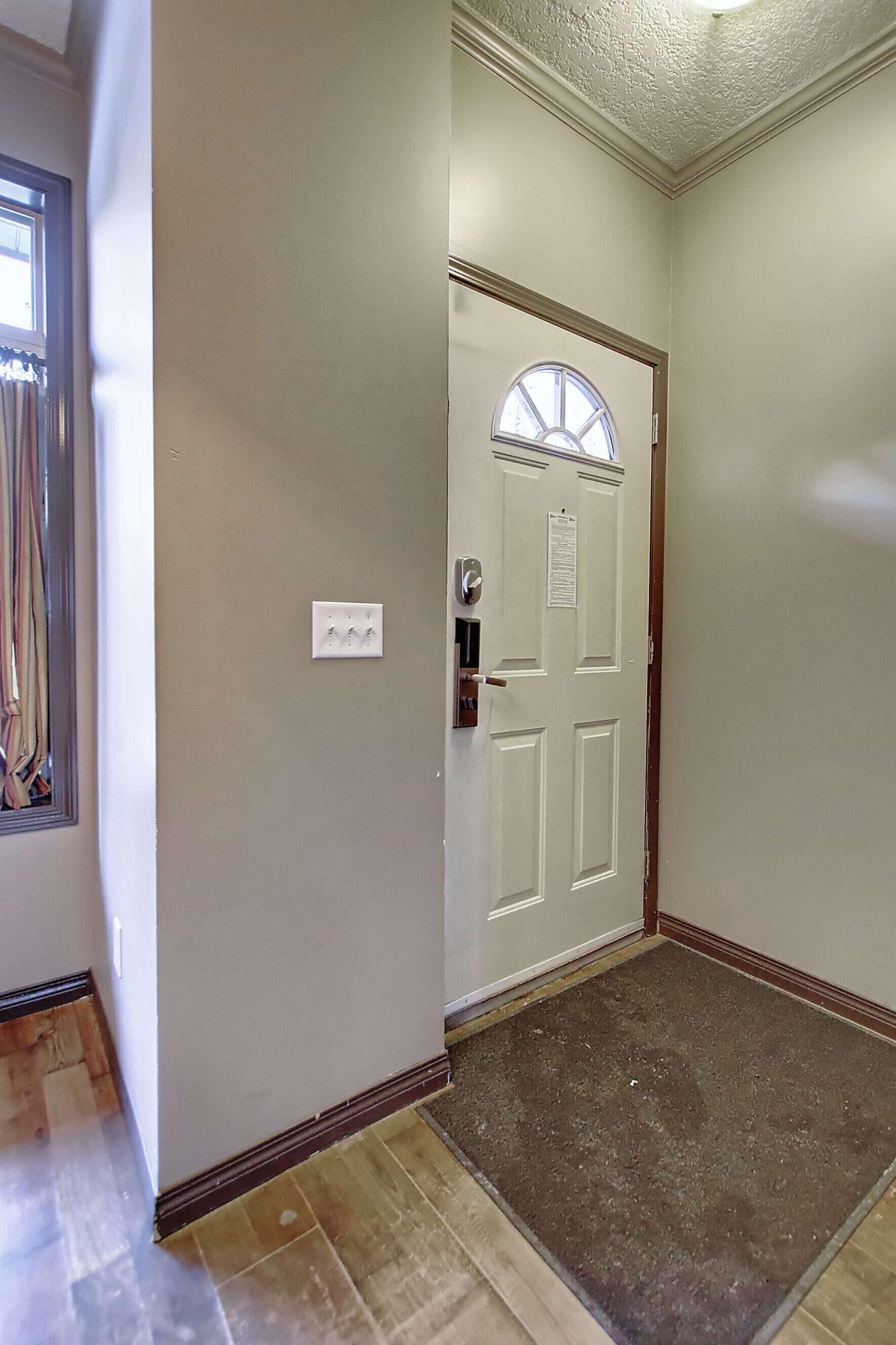 A hallway with a white door, a small window with curtains, and a light switch on the wall.